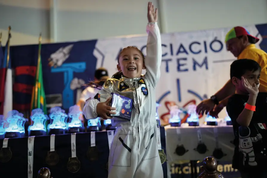Niña vestida con traje de astronauta levantando la mano con entusiasmo mientras sostiene un premio, frente a una mesa con trofeos iluminados y un fondo con temática STEM y espacial.