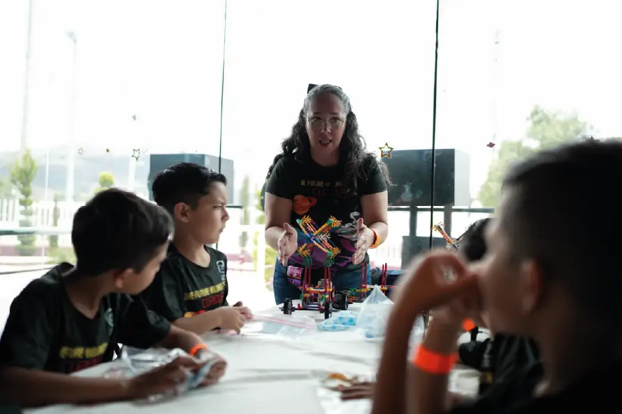 Instructora explicando un proyecto STEM a un grupo de niños alrededor de una mesa con estructuras y materiales de construcción, en un espacio iluminado con ventanales al fondo.