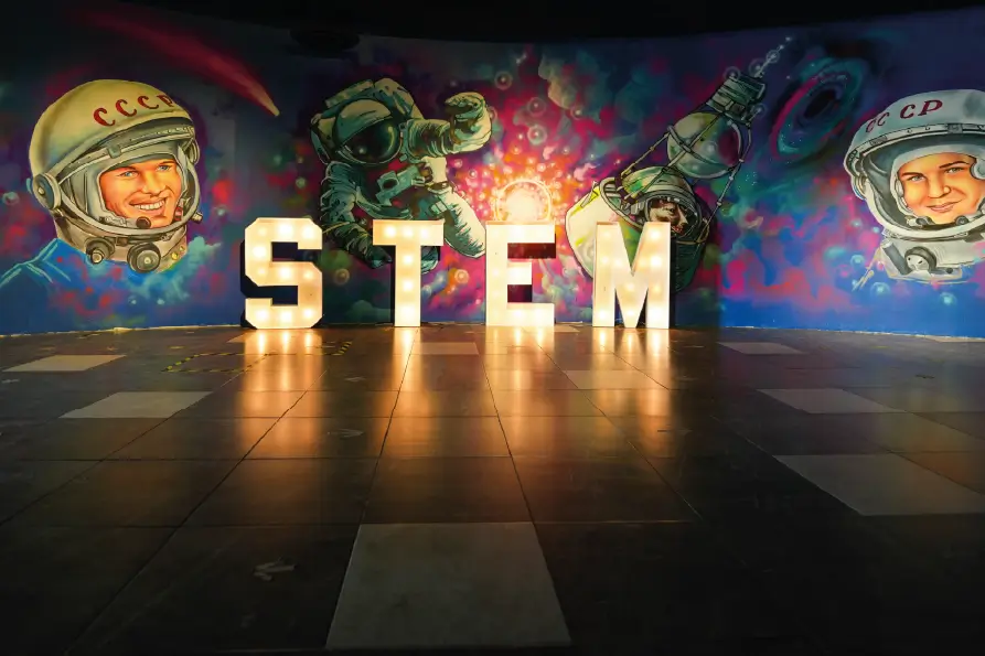 Letras luminosas que forman la palabra “STEM” frente a un mural colorido con ilustraciones de astronautas y elementos espaciales, en un espacio interior con piso de baldosas.